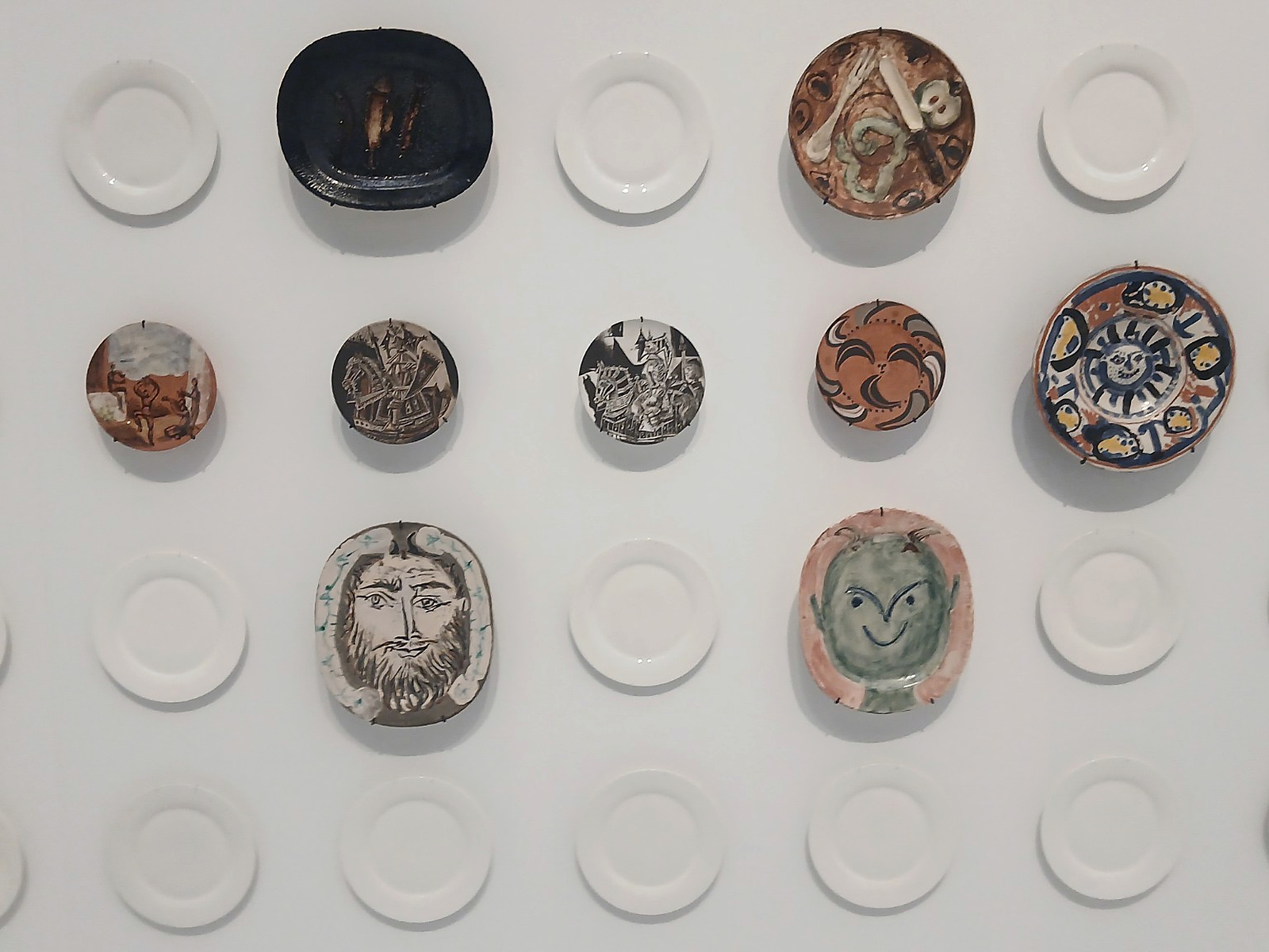 A gallery wall covered with ceramic plates, including painted faces and patterns among rows of white plates.